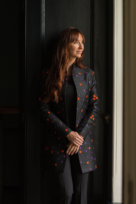 Woman leaning against wall by a window in a dark silk Bhumi jacket with floral embroidery