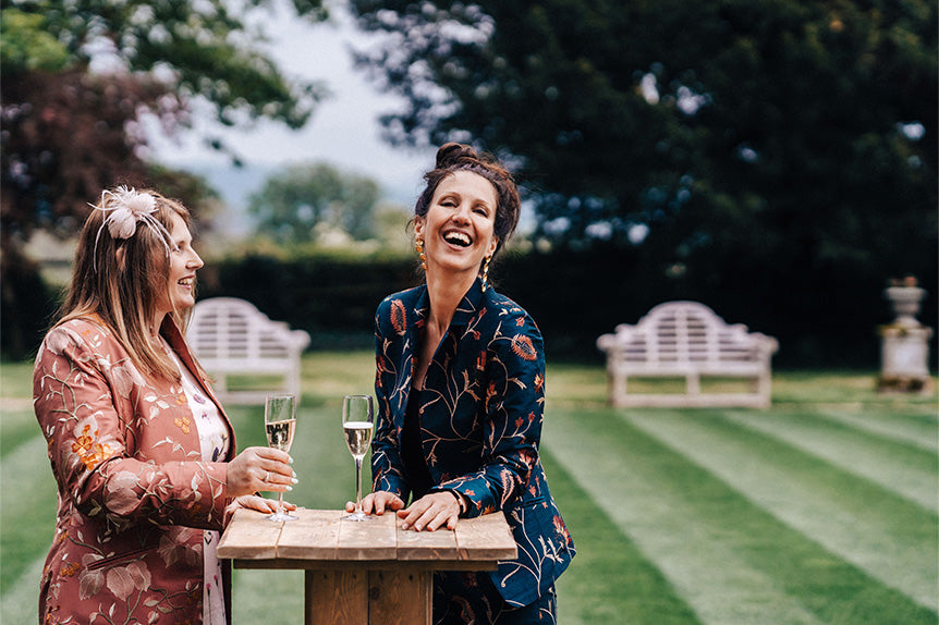 Two women laugh while drinking champagne at a wedding wearing silk coats