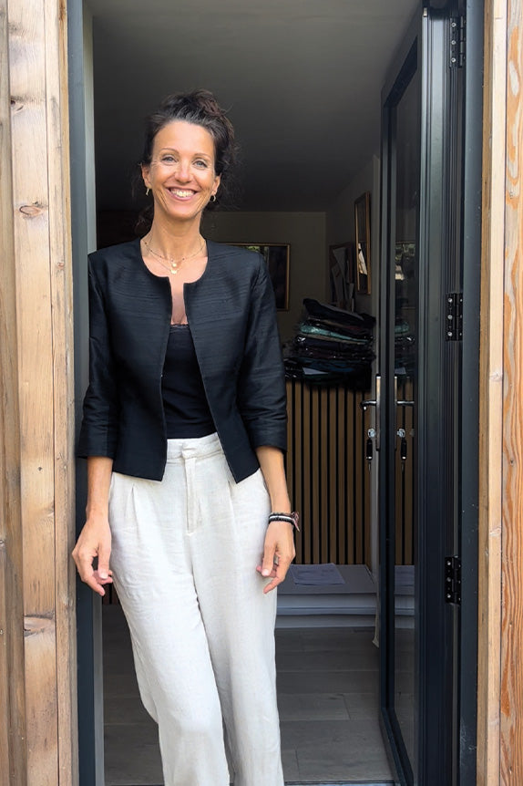 Woman with dark hair stands in office door wearing light linen trousers and a cropped black silk Juna jacket