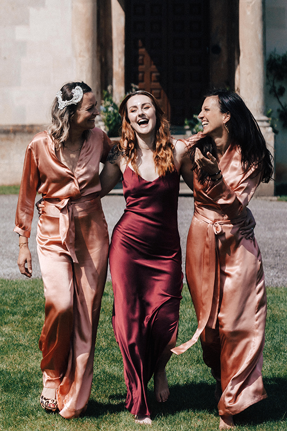 three women, bridesmaids, walking toward the camera laughing on a sunny day in front of a stately home, one woman in a cherry red silk slip dress, two women in blush pink silk jumpsuits.