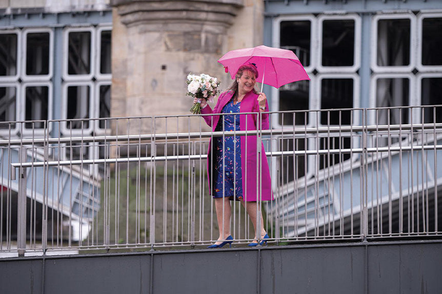 Woman stands on bridge on her way to be a wedding guest in a bright pink coat and blue dress, she wears a lyra coat in wild orchid and a marilyn dress in african cobalt