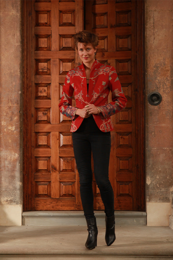 Woman with short brown hair walking down steps in front of a large wooden front door in black jeans and a tailored red silk Anya jacket with embroidery detail