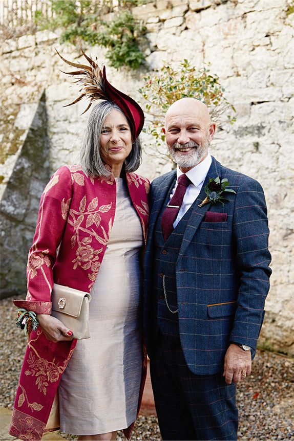 Mother and father of the bride, mother of the bride in pale pink dress with shibumi lyra coat in deep raspberry cashmere with gold embroidery and matching red hat
