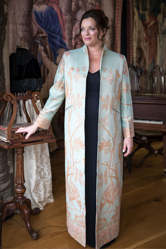 woman standing in ornate room next to a music stand and piano wearing a black maxi dress and a pale blue embroidered floor length opera coat