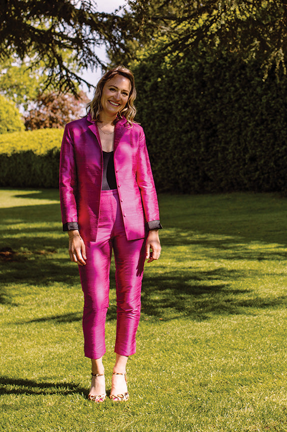 blonde woman standing on grass under a tree in a bright pink silk suit. shibumi nina jacket and cigarette trouser in hot pink