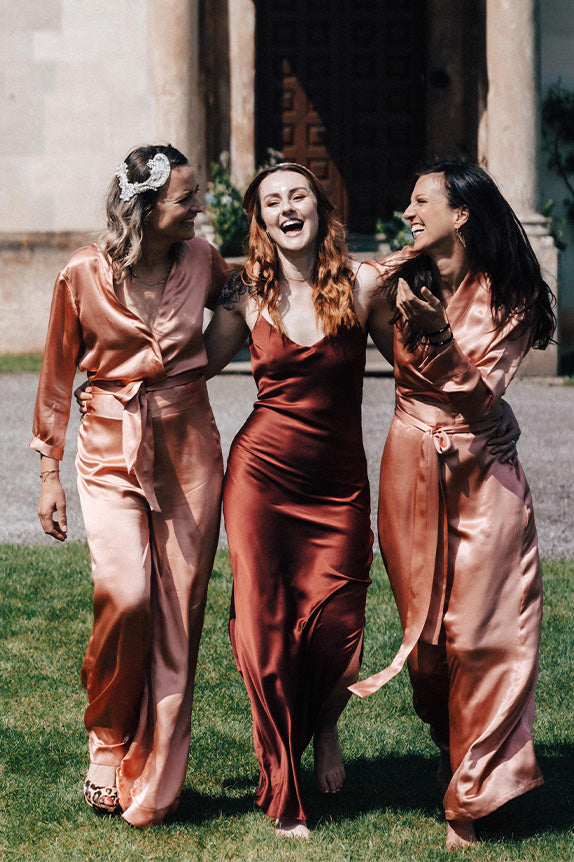 Three women walking and laughing outside wearing satin silk bridesmaids slip dress and blush rose satin silk bridesmaids jumpsuit
