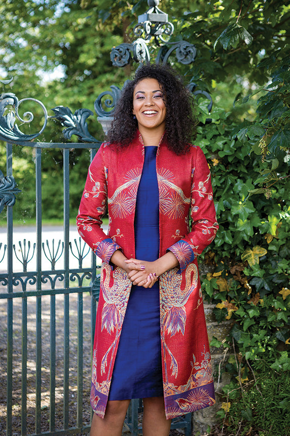 Woman in garden wearing colourful cashmere coat over blue dress. Lyra coat in Venetian Red.