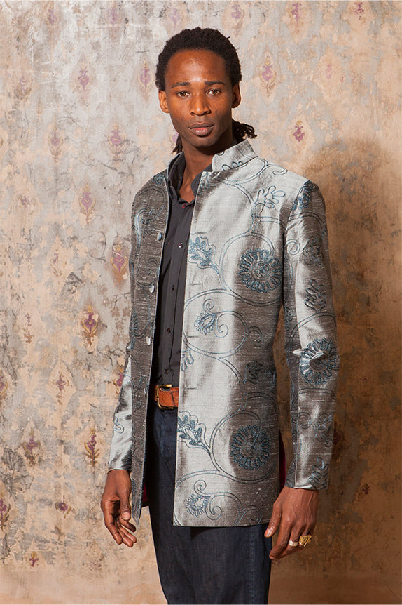 black man standing in front of wall ornate wall paper wearing a shirt, trousers and a silver and blue nehru jacket