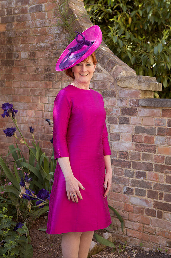 redhead woman standing in garden in front of brick wall wearing bright pink dress with long sleeves and a pink hat