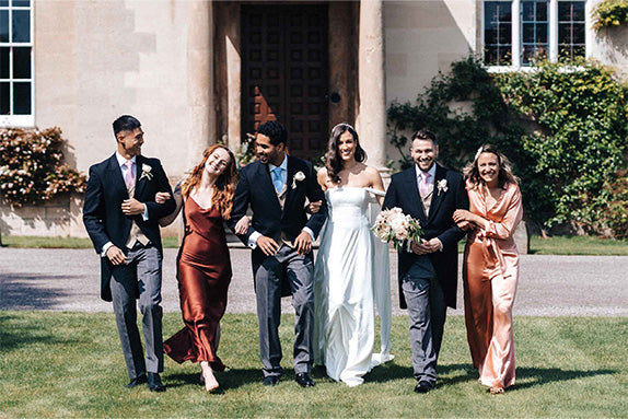 Wedding party laughing walking toward camera across grass in front of a stately home with men wearing grey and black tailsuits with pink and blue ties, bride in off shoulder white dress, bridesmaids in a cherry red silk slip dress and blush pink jumpsuit.
