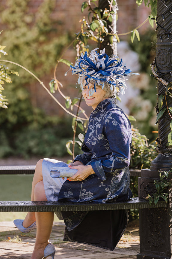 Woman sitting on bench surrounded by flowers in a dark blue coat, pale blue shift dress and blue fascinator hat holding a pale blue silk clutch bag.