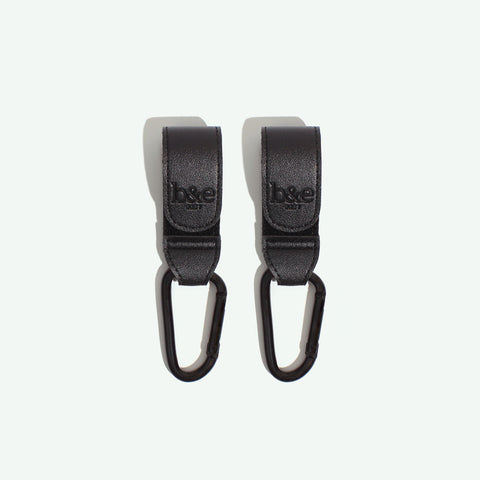 A pair of black adjustable pram straps made from vegan leather and heavy duty velcro. The straps have black metal hardware and metal spring-lock hooks.