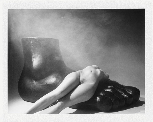 Black and white photo of a nude woman laying on a large black shoe.
