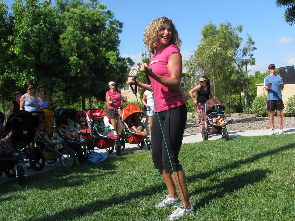 Lisa leading a class of Mom's through a set of exercises with a resistance band. She's smiling and wearing workout gear and the other moms have their strollers with them.