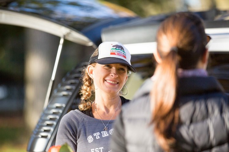 Lisa wears a baseball cap standing next to a minivan, smiling and talking to another woman with a ponytail whose back is to the camera.