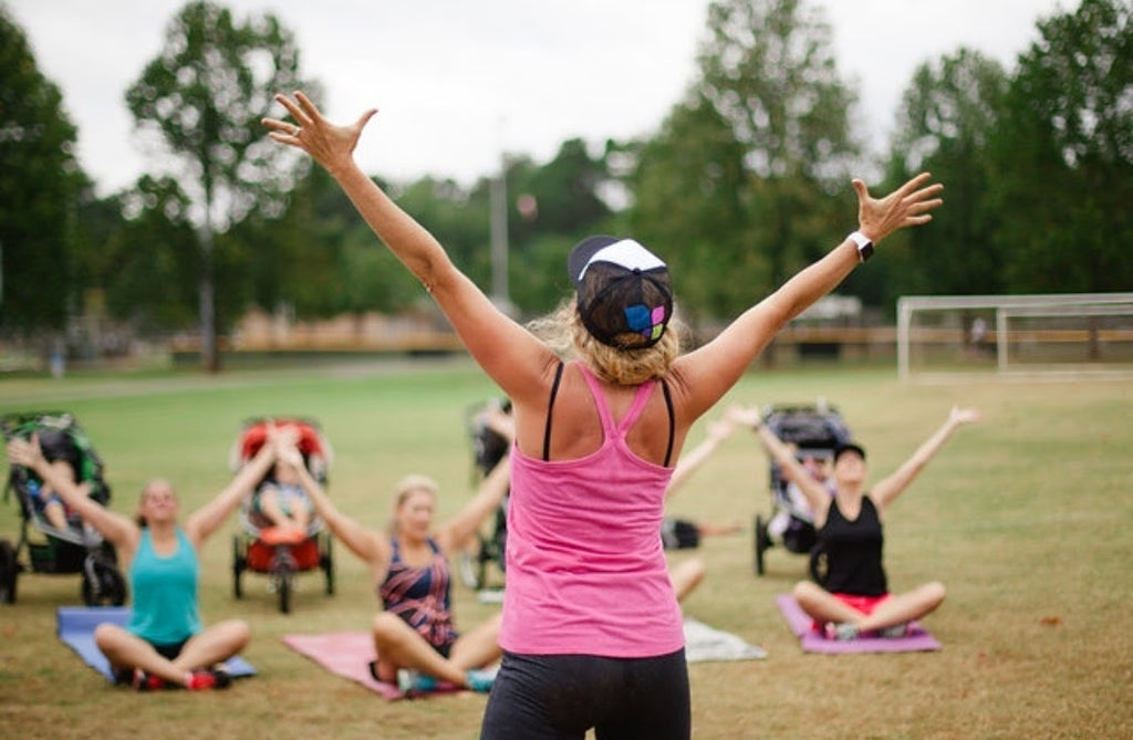 Lisa holds her arms out above her while teaching a class. She's wearing a pink tanktop and sports bra, and has her hair up in a baseball cap. Moms sit on yoga mats in front of her, mimicking her movement and stretching.