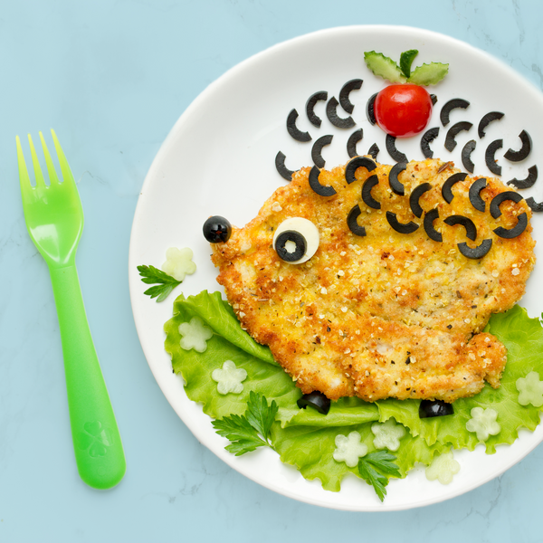 Image of a plate of food next to a bright green fork. On the plate is a piece of chicken schnitzel decorated to look like a hedgehog on top of a bed of lettuce as grass with cut cheese flowers. The hedgehog's spines are made of sliced olives and it has a cute face and small feet also made of olives, with a cherry tomato on top.