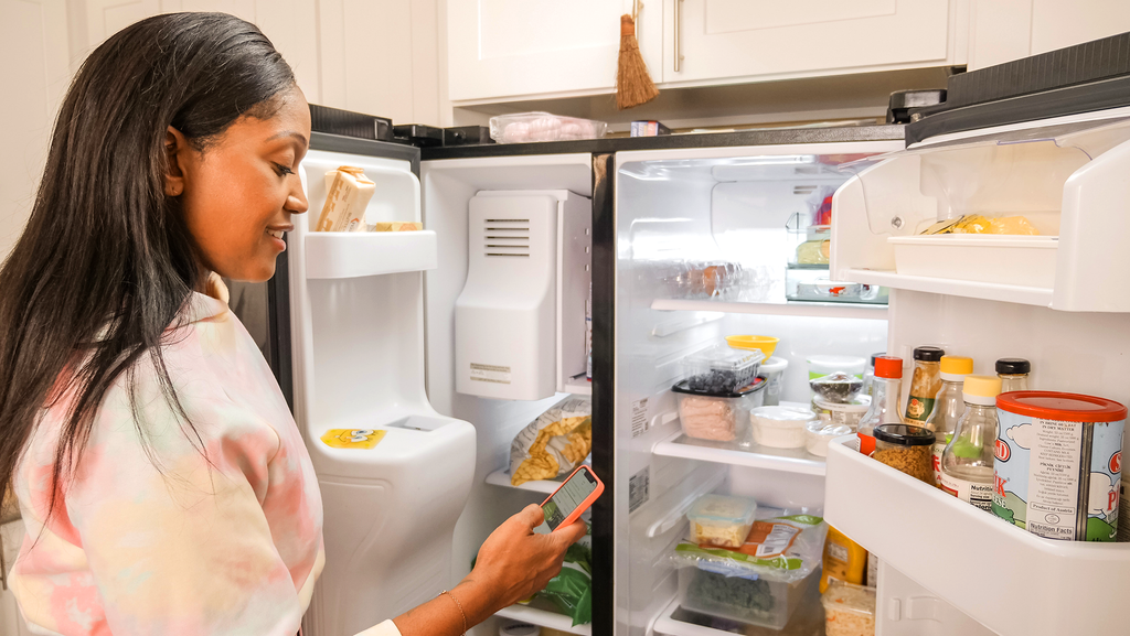 Image of a woman checking her phone while looking in her freezer and fridge, checking to see what ingredients she has available.