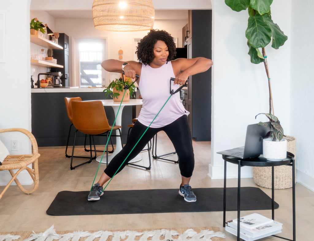 Image of a Mom in her living room in front of a laptop, doing stretches on a yoga mat holding a resistance band.