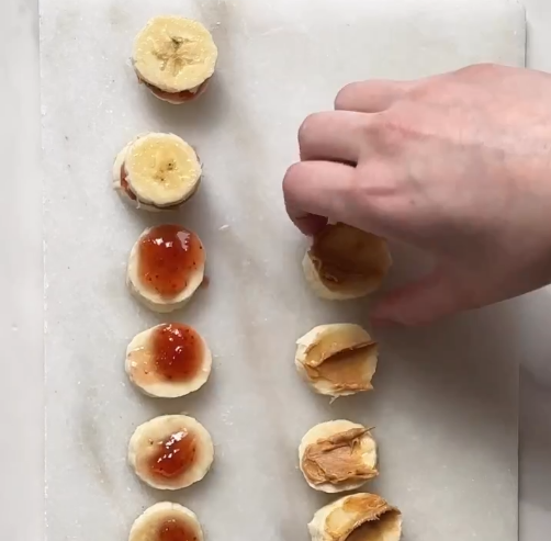 Overhead image of a hand placing slices of bananas on top of other slices that have been covered in peanut butter and jelly, making small pseudo sandwiches. Image from @reallittlemeals on instagram