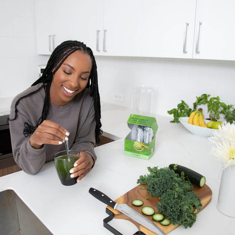 Black woman holding a cup of green juice on the kitchen counter next to Fine USA's Green Barley and Chia Seed product