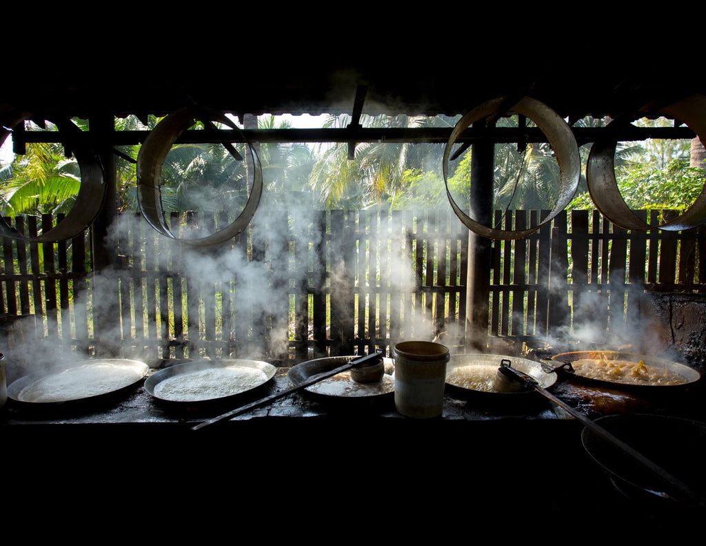 Beneath a thatched roof, steam rises from a line of shallow vats containing bubbling coconut sap.