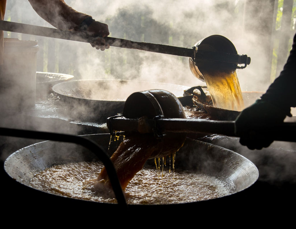 Two people use ladles to pour boiling coconut sap into large, shallow bowls.