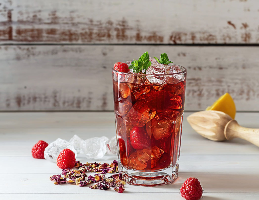 A glass of ice raspberry red juice surrounded by fresh raspberries, ice cubes, and half a lemon.