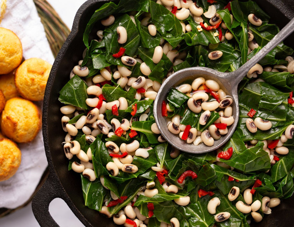 A skillet filled with vibrant collard greens, diced red peppers, and black eye peas. A plate of cornbread sits to one side.