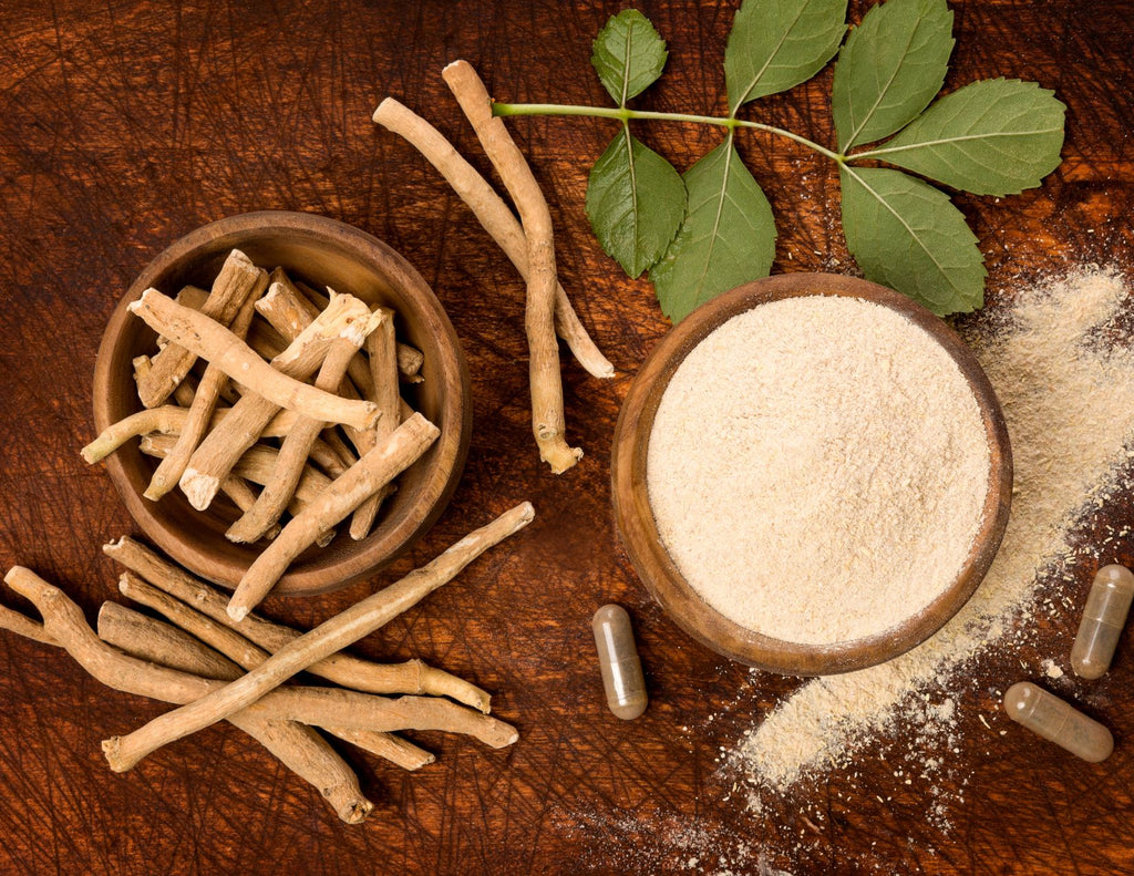 The light brown powder, roots, and green leaves of ashwagandha on a dark brown cutting board.