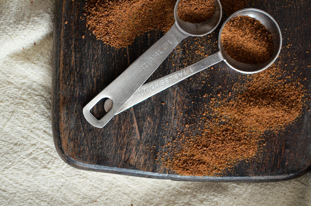 Two metal measuring spoons filled with coconut sugar rest on a dark wood cutting board.