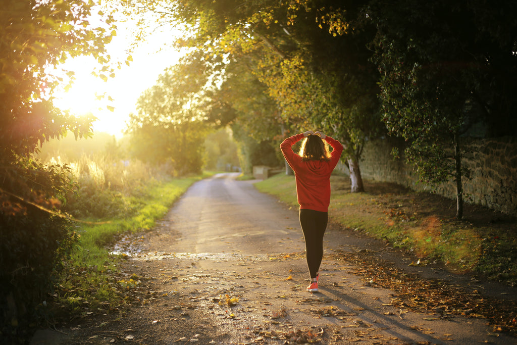 A woman with her hand on her head walks along a wood path with sunlight streaming through the trees.