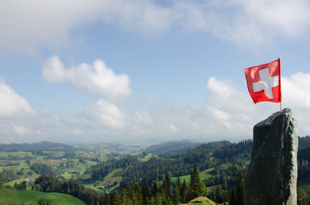 A Swiss flag, red with a white cross, sits atop a rock jutting from a mountain top.