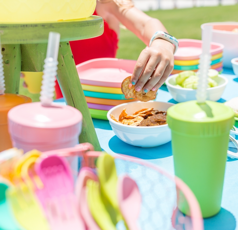 A range of different colour re-play bowls and re-play cutlery and re-play cups on a blue tablecloth with a childs hand stealing some treats from a bowl