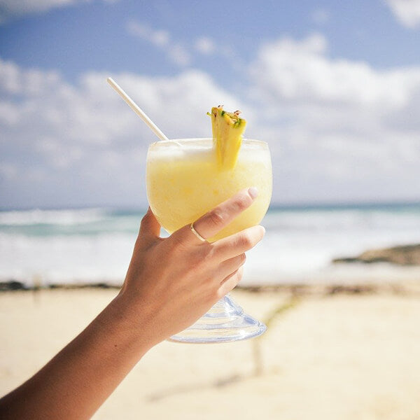 pina colada cocktail am Strand in der Hand einer Frau mit Wasser im Hintergrund