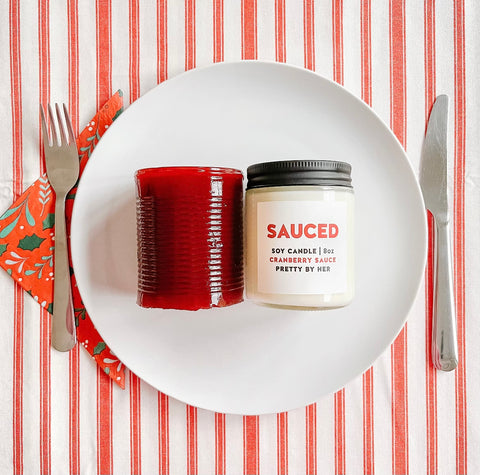 A candle that reads "Sauced" and is scented like Cranberry Sauce sits on a Thanksgiving Dinner Plate next to canned cranberry sauce.