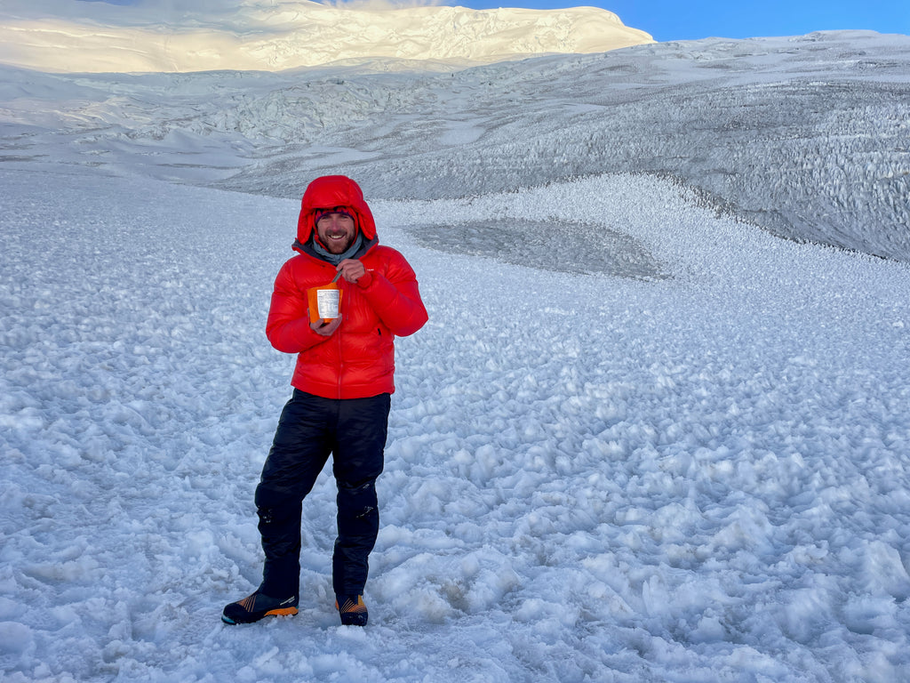 A man wearing a bright red jacket and other climbing clothes stands in a snowfield  eating an Expedition Foods meal pouch.