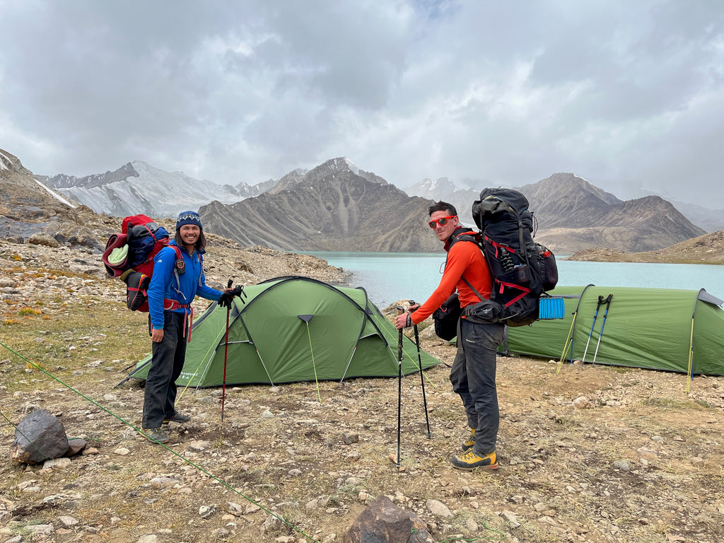 Two men with very large expedition rucksacks on their backs stand outside a green tent. The tent is pitched on a rough grassy spot. In the back ground we can see milky blue glacial lakes and tall rocky mountains.