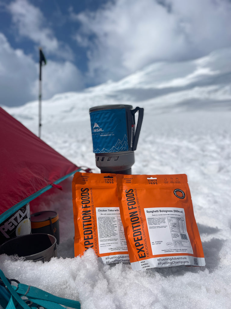 Outside a red tent pitched on a field of snow is a small camping stove. Next to the stove are two Expedition Foods meal pouches.