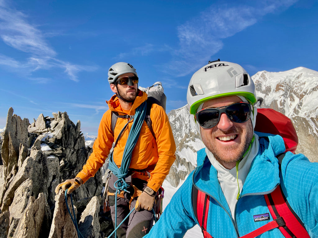 Two men face the camera smiling. they are on top of a jagged mountain surrounded by mountains and blue sky.