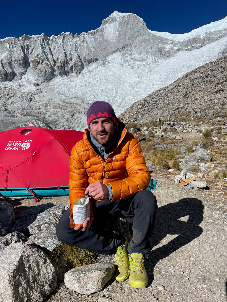 A man in an orange down jacket crouches down outside a tent holding a pouch of Expedition Foods freeze-dried food. he's eating it with a spoon. In the background he's surrounded by high snowy mountains.