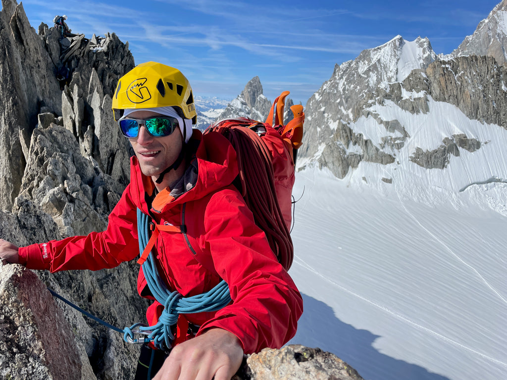 A climber wearing a bright yellow helmet and red jacket is on a jagged rock ridge. In the background are more jagged rocky mountains and snow slopes. The sky is bluebird blue.