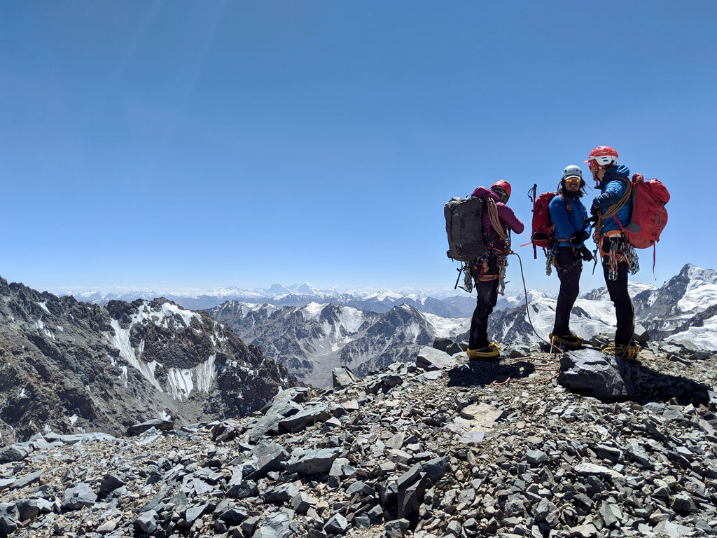 Three men dressed for the mountains and wearing climbing equipment stand on top of a mountain. They are surrounded by beautiful blue skies and mountains in all directions.