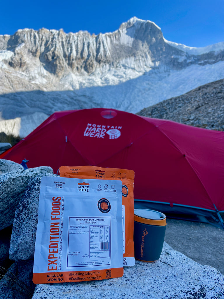 Blue skies and high snowy mountain surround a bright red tent pitched on rocky terrain. In the foreground are two Expedition Foods meal pouches waiting to me prepared for someone's dinner