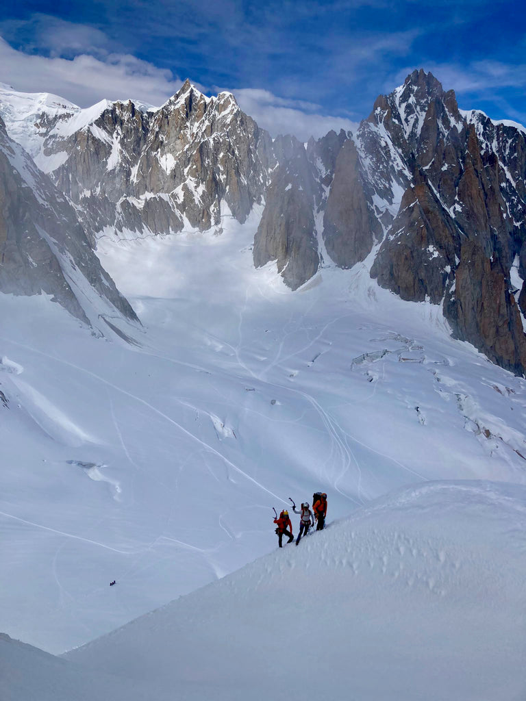 Blue skies and rocky snowy mountains dwarf three small figures on a steep snow slope.