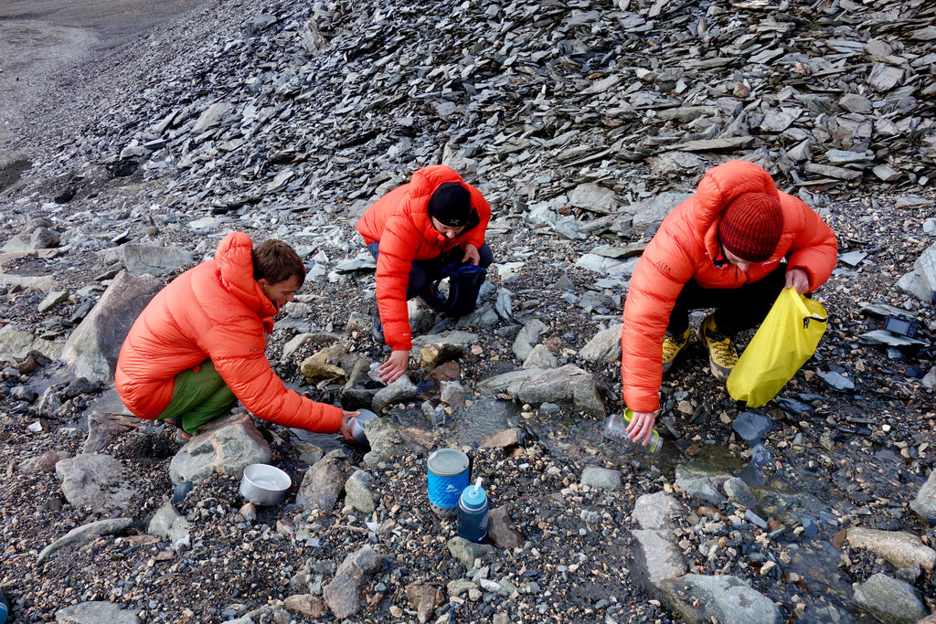 The people wearing matching orange down jackets crouch down next to a tiny trickling stream. They are scooping water into their water bottles and larger containers.