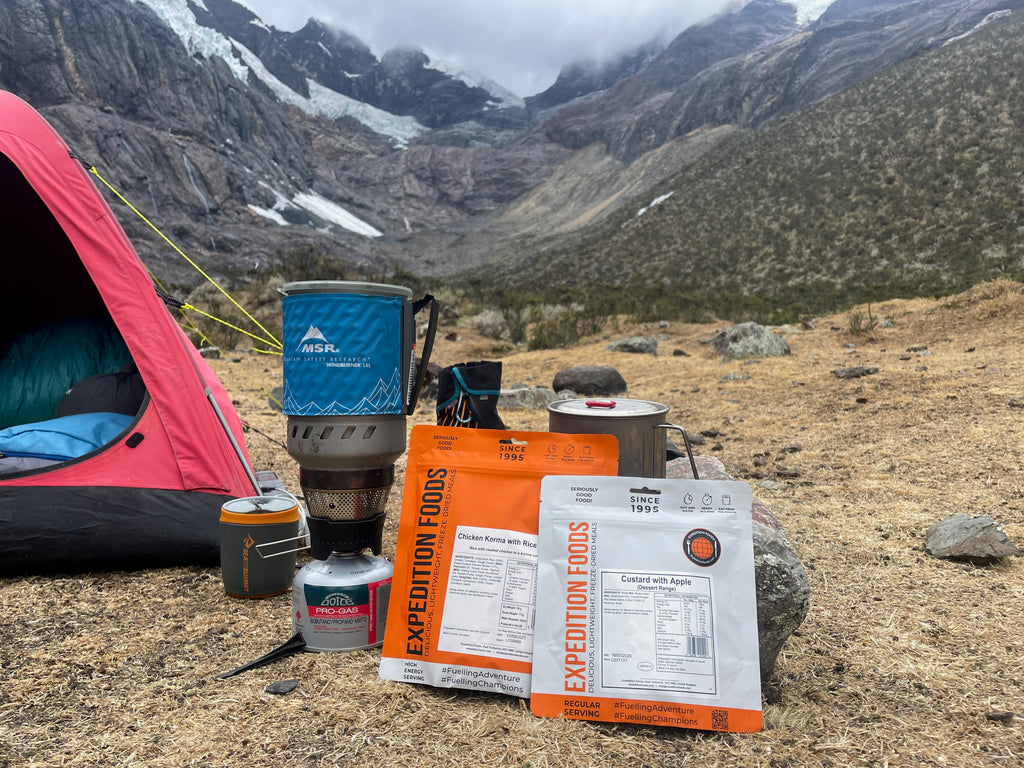 Steep grassy slopes rise up into rocky mountains. In the foreground a bright red tent is pitched. A small camping stove is set up and in front of it are two Expedition Foods meals waiting to be prepared for someone's dinner