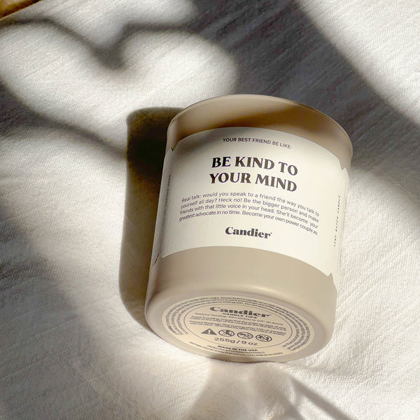 A warm gray candle with a white label that reads Be Kind To Your Mind