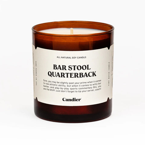 An amber colored scented candle with a cream label that reads Bar Stool Quarterback