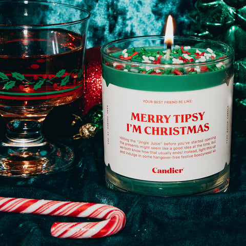 A Holiday themed scented candle with a label that reads merry tipsy I'm christmas